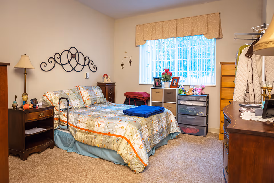 A cozy bedroom with a bed covered in floral bedding and a blue blanket folded on top. There is a wooden nightstand with a lamp and small items beside the bed. The room has a window with vertical blinds and a beige valance, letting in natural light. Below the window are storage units with framed photos, a vase with flowers, and stuffed animals. The room also features a wooden dresser, a coat rack with a beige jacket, and beige carpeted flooring.