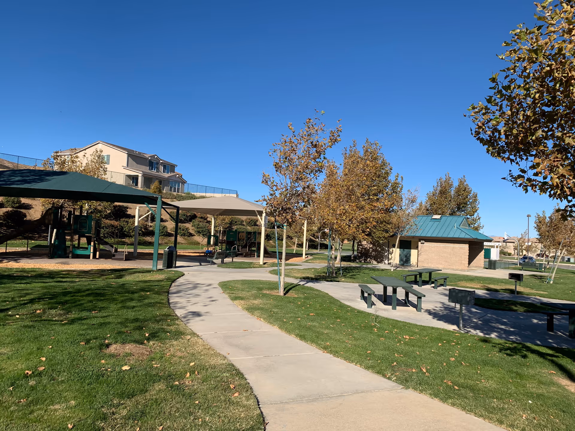 A sunny outdoor park area with a paved walkway, green grass, several trees with autumn-colored leaves, picnic tables with attached benches, a small building with a green roof, and shaded playground equipment in the background.