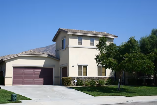 Two-story beige house with an attached two-car garage, driveway, and front lawn under a clear blue sky.