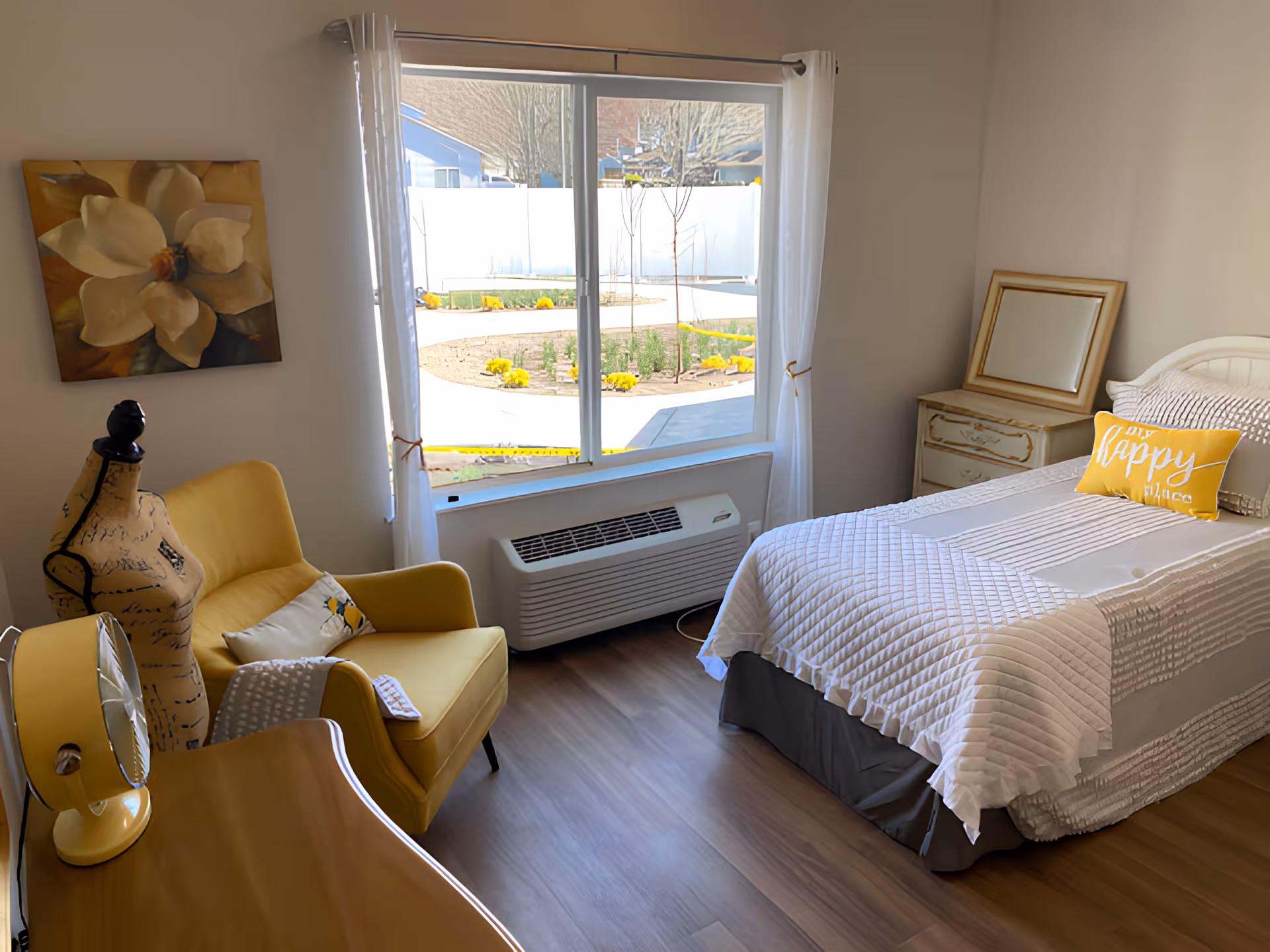 A cozy bedroom with a single bed covered in a white quilt and a yellow pillow that says 'my happy place'. There is a yellow armchair with a decorative pillow and a mannequin next to it. A wooden desk with a yellow lamp is in the foreground. A large window with white curtains shows a view of a garden with yellow flowers outside. A small dresser with a framed mirror is beside the bed.