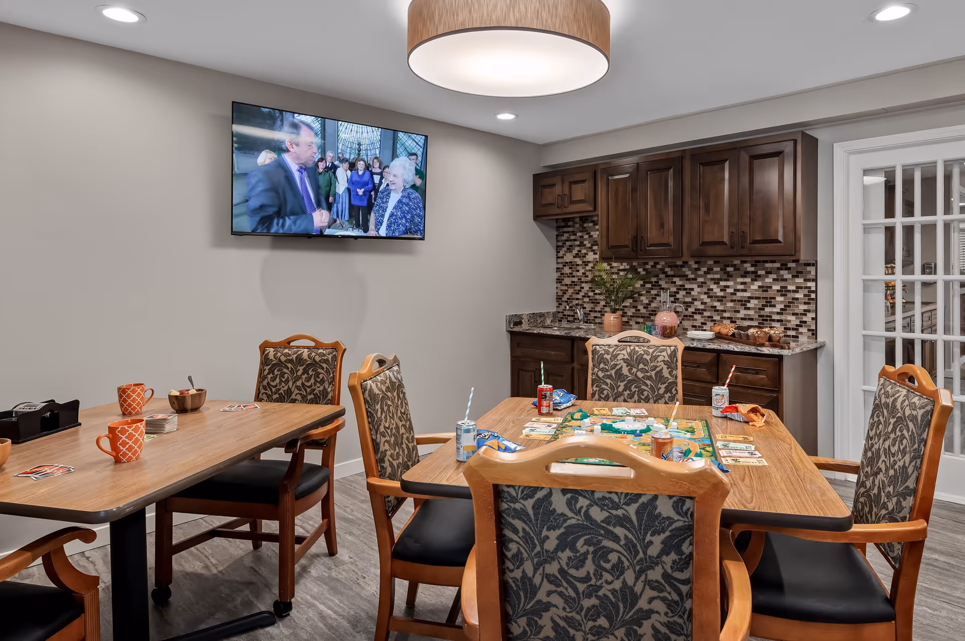 A cozy common room with two wooden tables and six upholstered chairs. One table has a board game set up with snacks and drinks, while the other has coffee mugs and playing cards. A flat-screen TV is mounted on the wall showing a group of people. The room features a small kitchenette with dark wood cabinets, a mosaic tile backsplash, and a granite countertop. A round ceiling light fixture illuminates the space.
