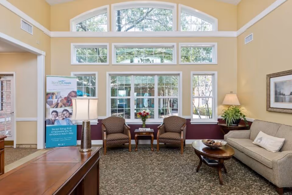 A cozy and well-lit living room area in Spring Creek Assisted Living with large windows showing outdoor greenery. The room features two patterned armchairs with a small wooden table and flower vase between them, a beige sofa with a pillow, a wooden coffee table with a decorative bowl, a side table with a lamp and plant, and a wooden desk with a lamp. A promotional banner for Spring Creek Assisted Living is visible near the entrance.