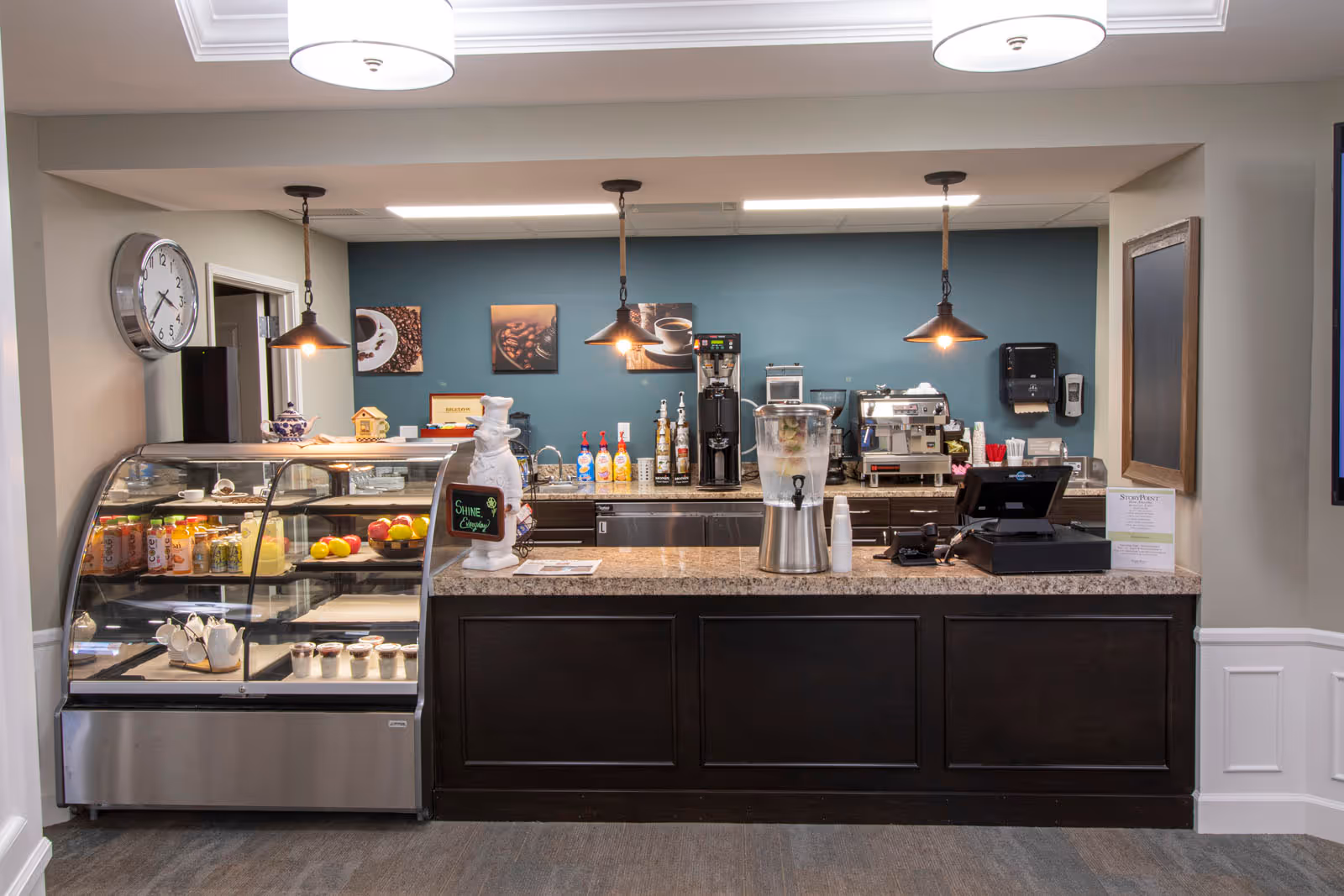 A coffee and snack bar area with a display case containing beverages and desserts, a counter with a water dispenser, coffee machines, syrup bottles, and a cash register. The wall behind is painted blue and decorated with coffee-themed pictures. There are three pendant lights hanging from the ceiling and a large clock on the left wall.