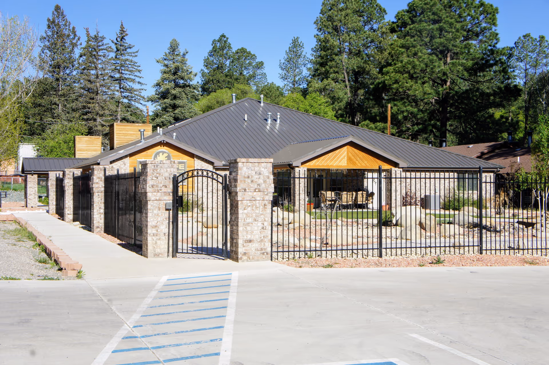 Exterior view of a single-story assisted living facility building with a dark metal roof, surrounded by a black metal fence with brick pillars. There is a paved walkway leading to a gated entrance, and outdoor seating is visible under a covered porch. Tall trees and greenery surround the property under a clear blue sky.