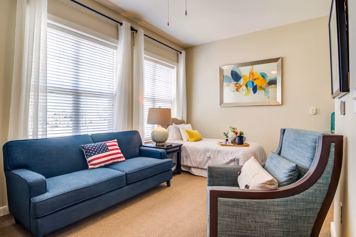 A cozy living space with a blue sofa featuring an American flag pillow, a blue armchair with a white pillow, a side table with a lamp, and a bed with white bedding and yellow accent pillows. The room has large windows with white blinds and sheer curtains, beige walls, and a colorful abstract painting above the bed.