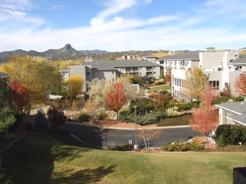 View of a senior living facility with multiple buildings surrounded by trees with autumn foliage, landscaped gardens, and a paved road. In the background, there are hills and a partly cloudy sky.