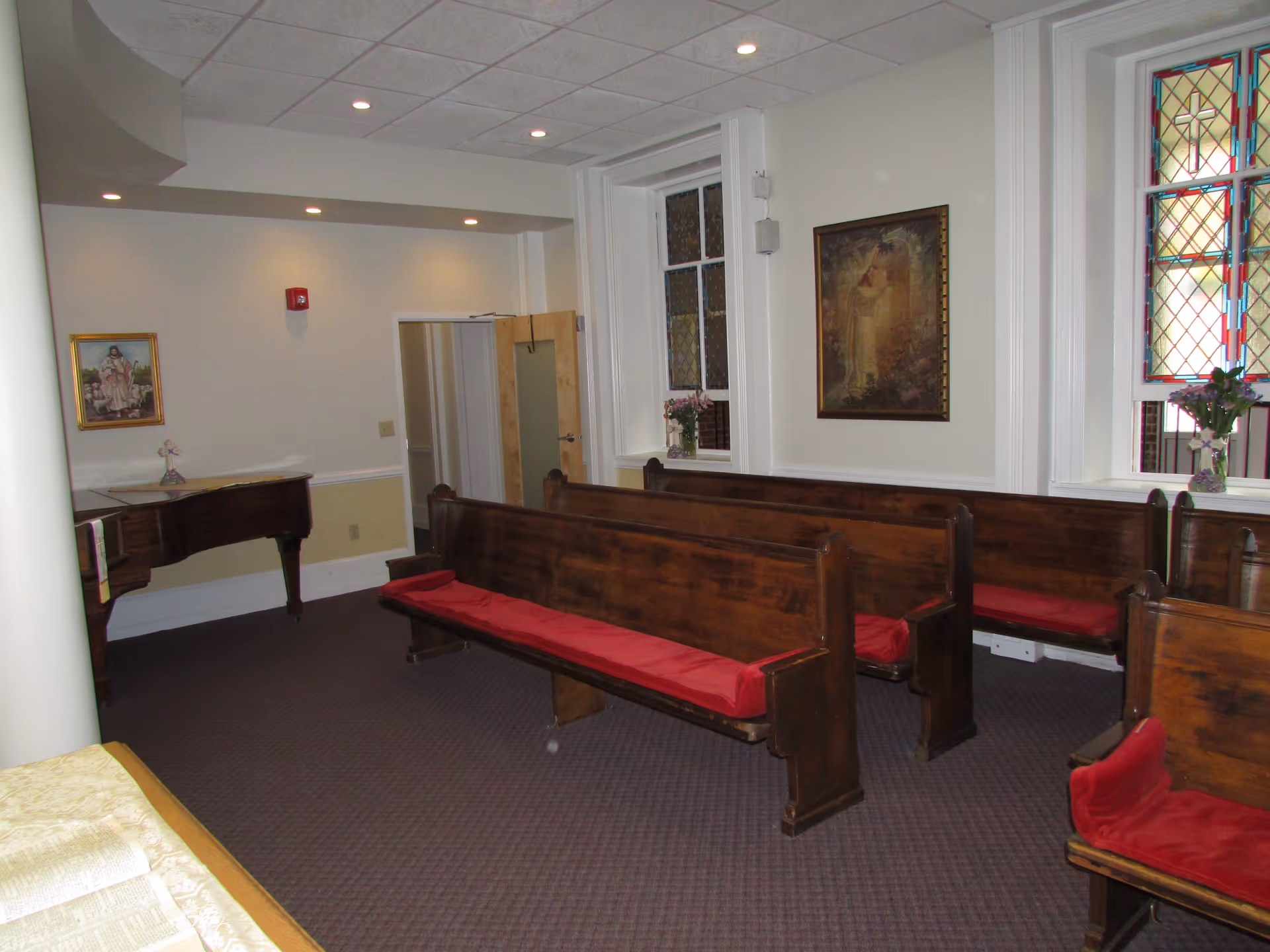 Interior room with wooden pews featuring red cushions, stained glass windows, religious paintings on the walls, and a grand piano in the corner.