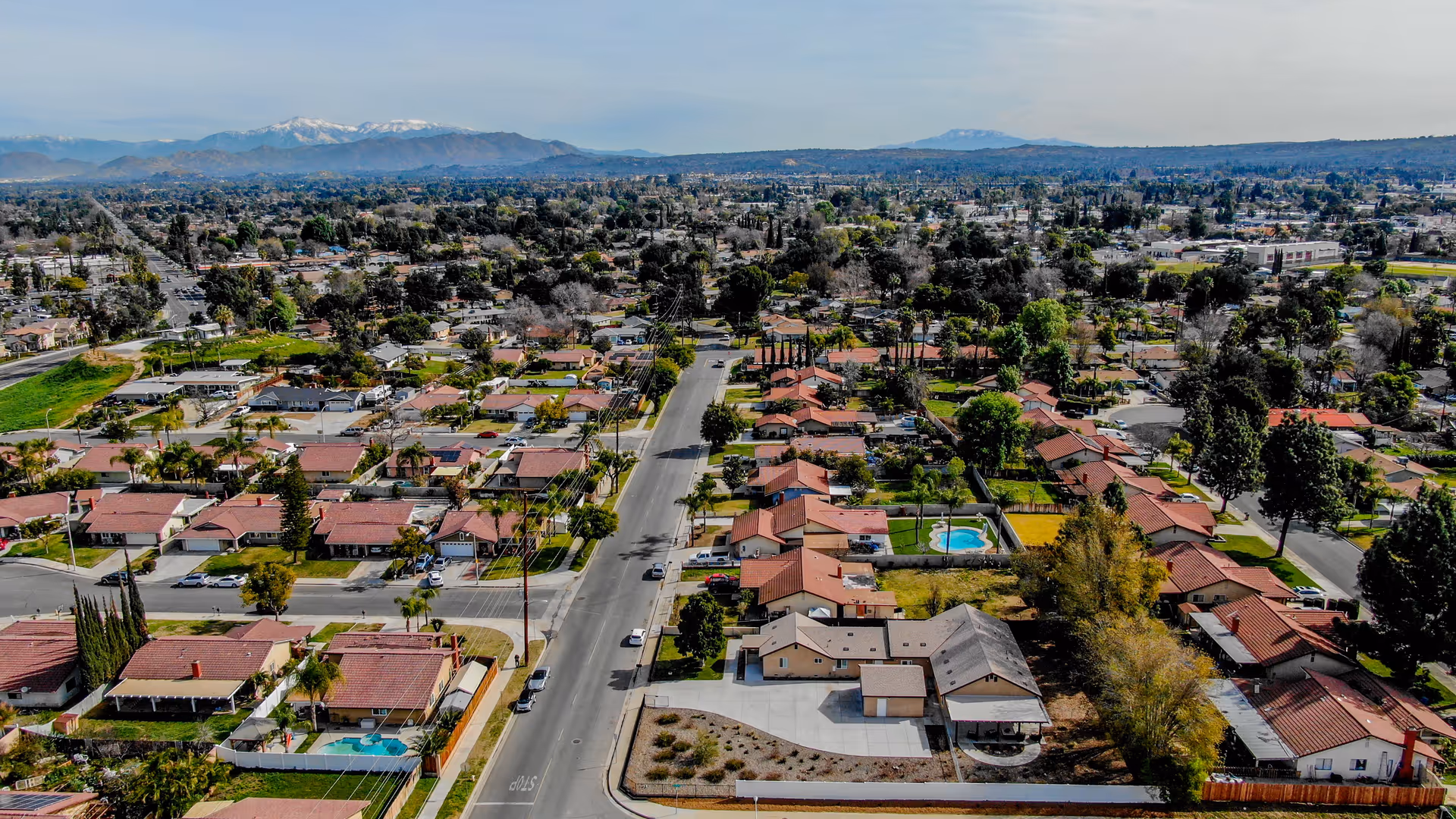 Aerial view of a suburban neighborhood with rows of houses featuring red-tiled roofs, streets lined with trees, and a few swimming pools visible in backyards. Mountains are visible in the background under a partly cloudy sky.