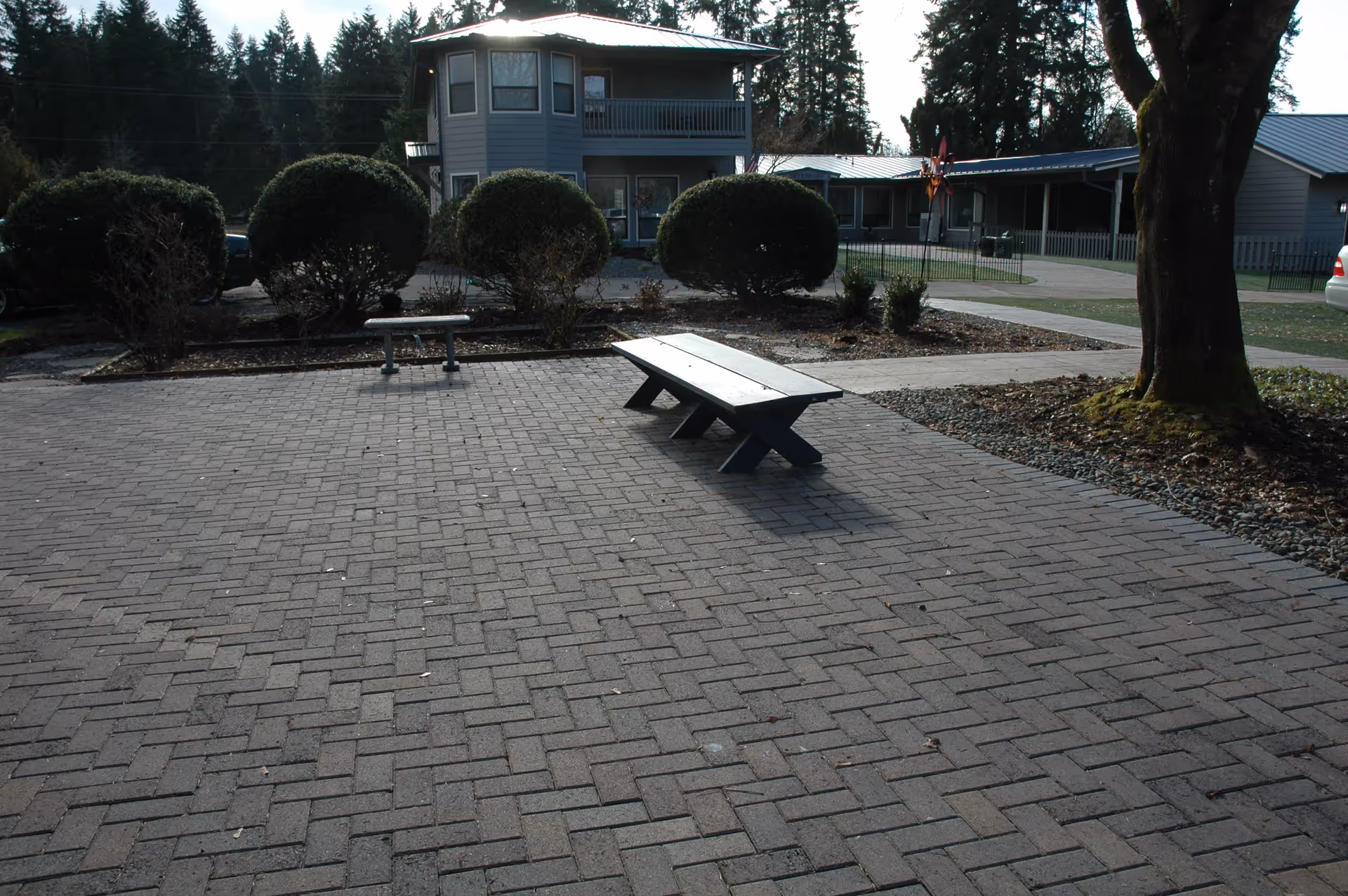 Paved courtyard with a low wooden bench, trimmed shrubs, and a two-story building in the background.