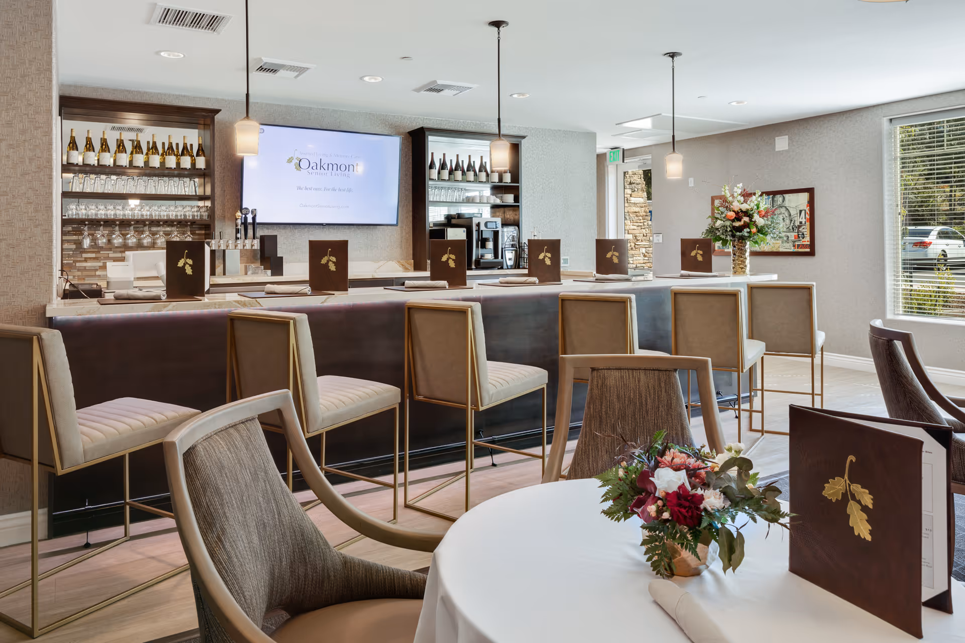A modern dining area with a bar counter featuring several high chairs. Behind the bar, shelves hold wine bottles and glasses, and a large screen displays the Oakmont Senior Living logo. A round table in the foreground is set with menus and a floral centerpiece. Large windows allow natural light to fill the room.
