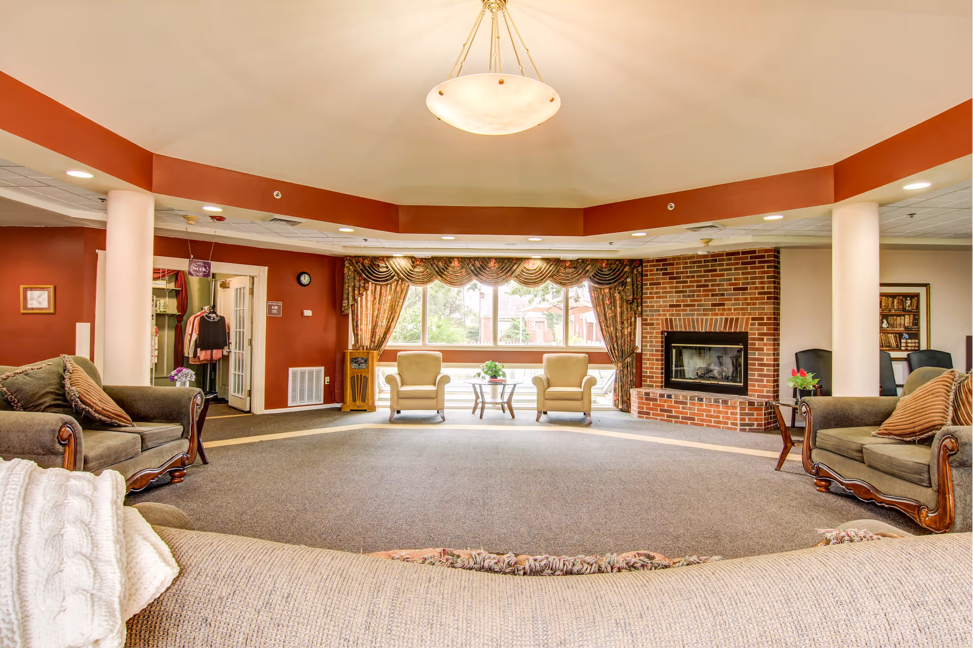 A spacious living room area with beige and brown sofas and armchairs arranged around a central carpeted floor. There is a brick fireplace on the right side and large windows with patterned curtains letting in natural light. The walls are painted a warm reddish-brown color, and there are white columns supporting the ceiling. A hanging light fixture is centered on the ceiling.
