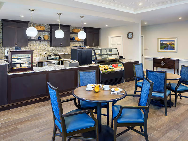 Dining area with round tables and blue chairs facing a service counter and refrigerated display case in a senior living facility café.