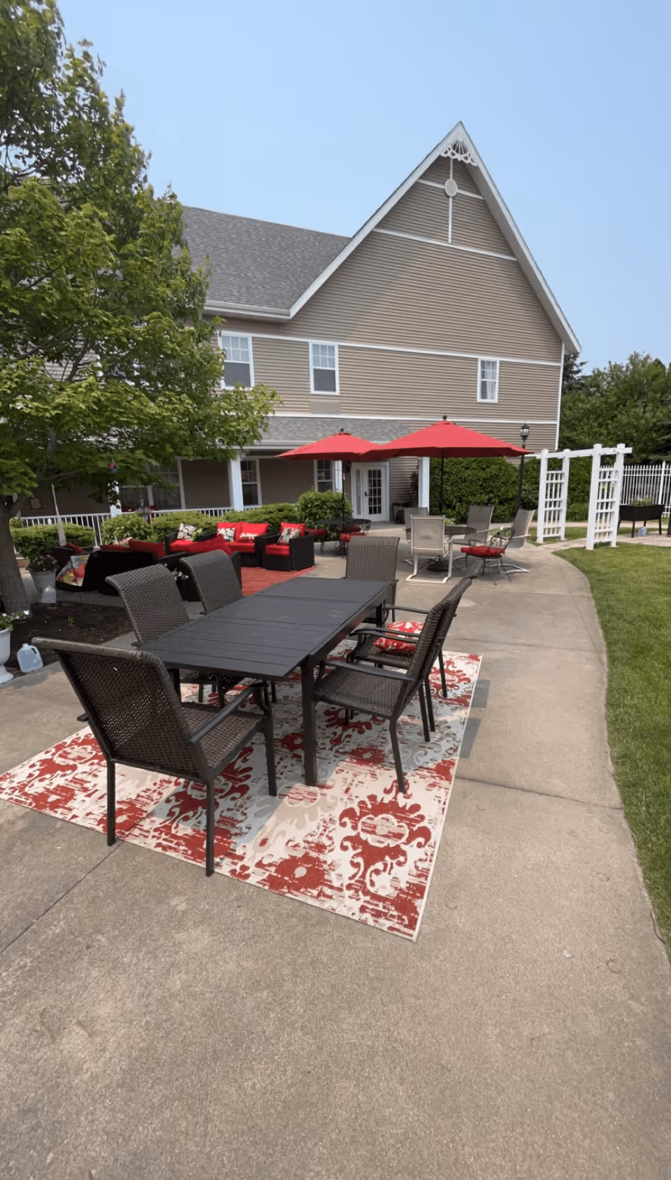 Outdoor patio area with a black rectangular table and six wicker chairs on a red and white patterned outdoor rug. In the background, there are red cushioned seating arrangements under red umbrellas, green bushes, and a large beige building with white trim. The area is surrounded by a concrete pathway and green grass.