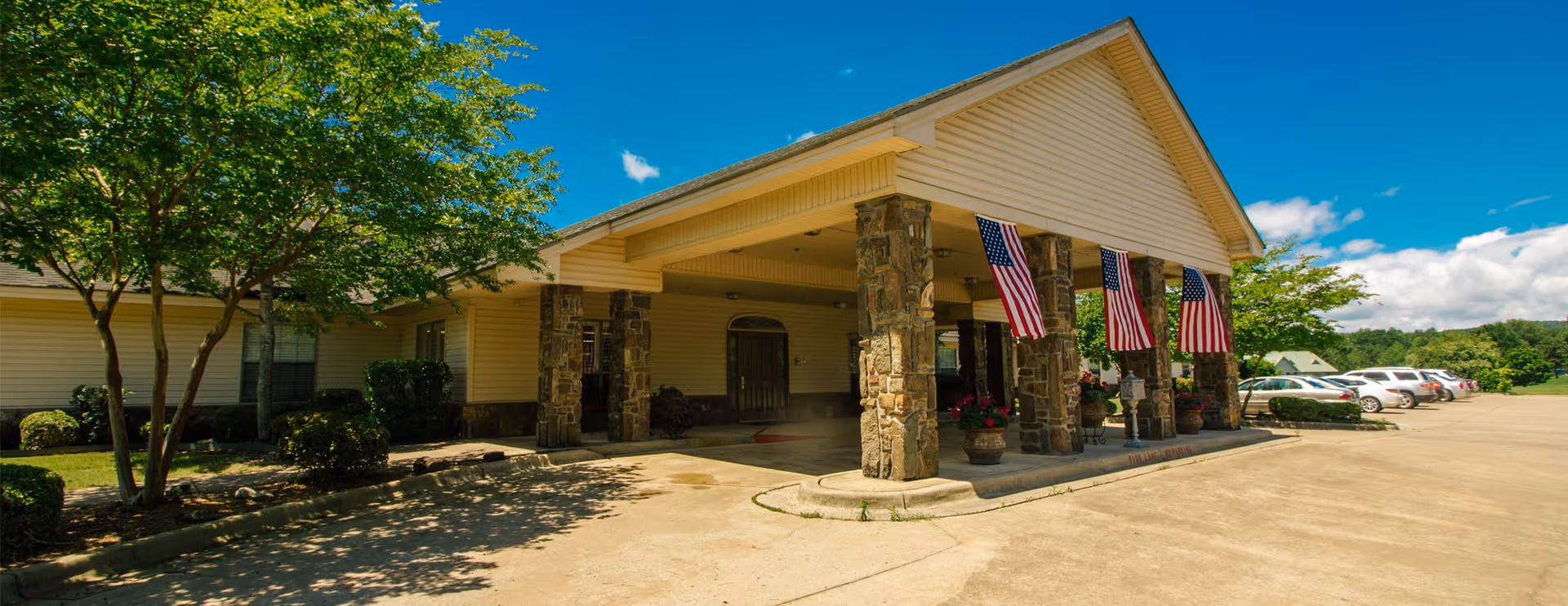 Front exterior view of Southridge Village Nursing and Rehabilitation Center featuring a covered entrance supported by stone pillars with American flags hanging from them, surrounded by trees and parked cars under a bright blue sky.