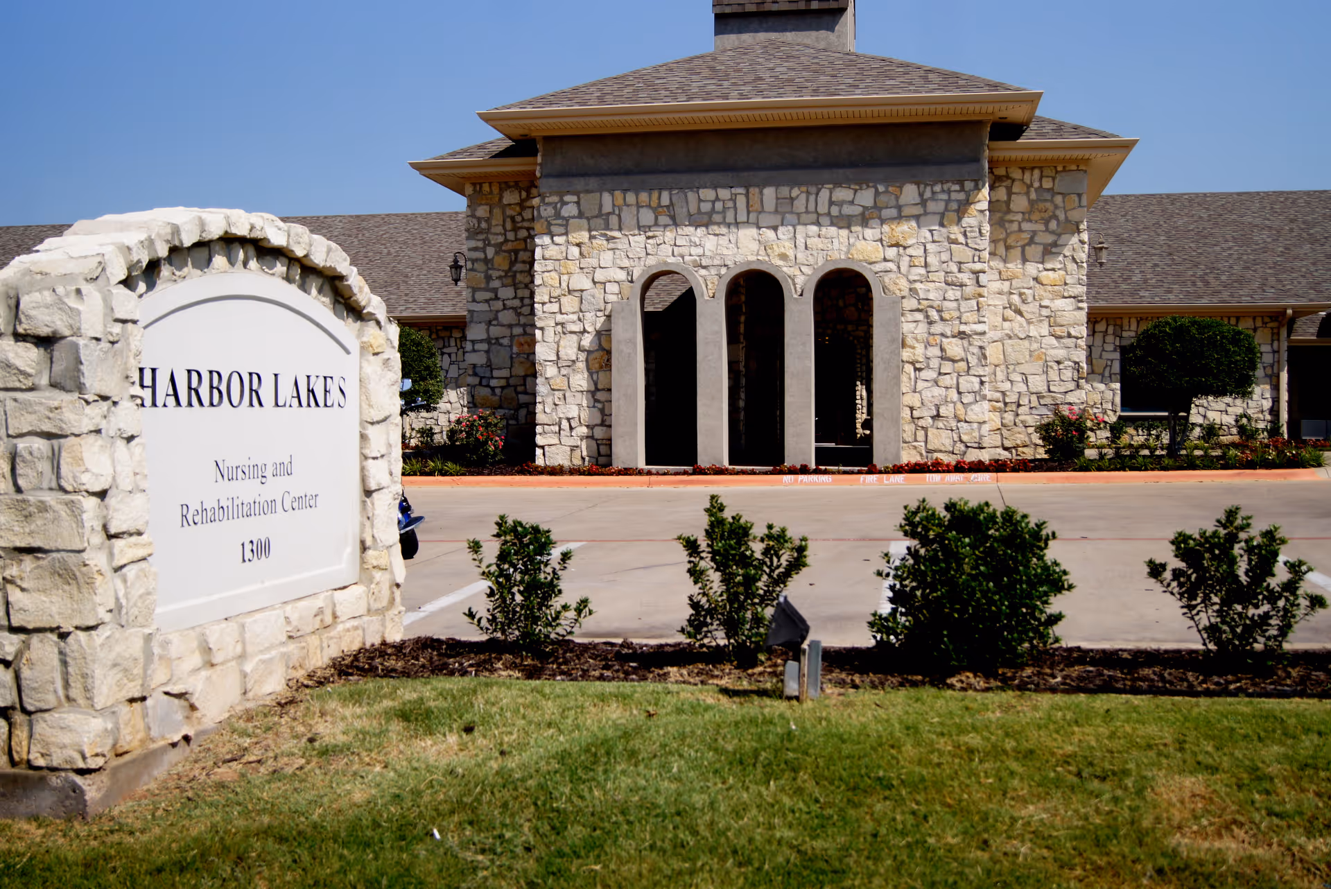 Stone sign for Harbor Lakes Nursing and Rehabilitation Center with the building entrance made of stone and three arched doorways in the background, surrounded by small bushes and a lawn.