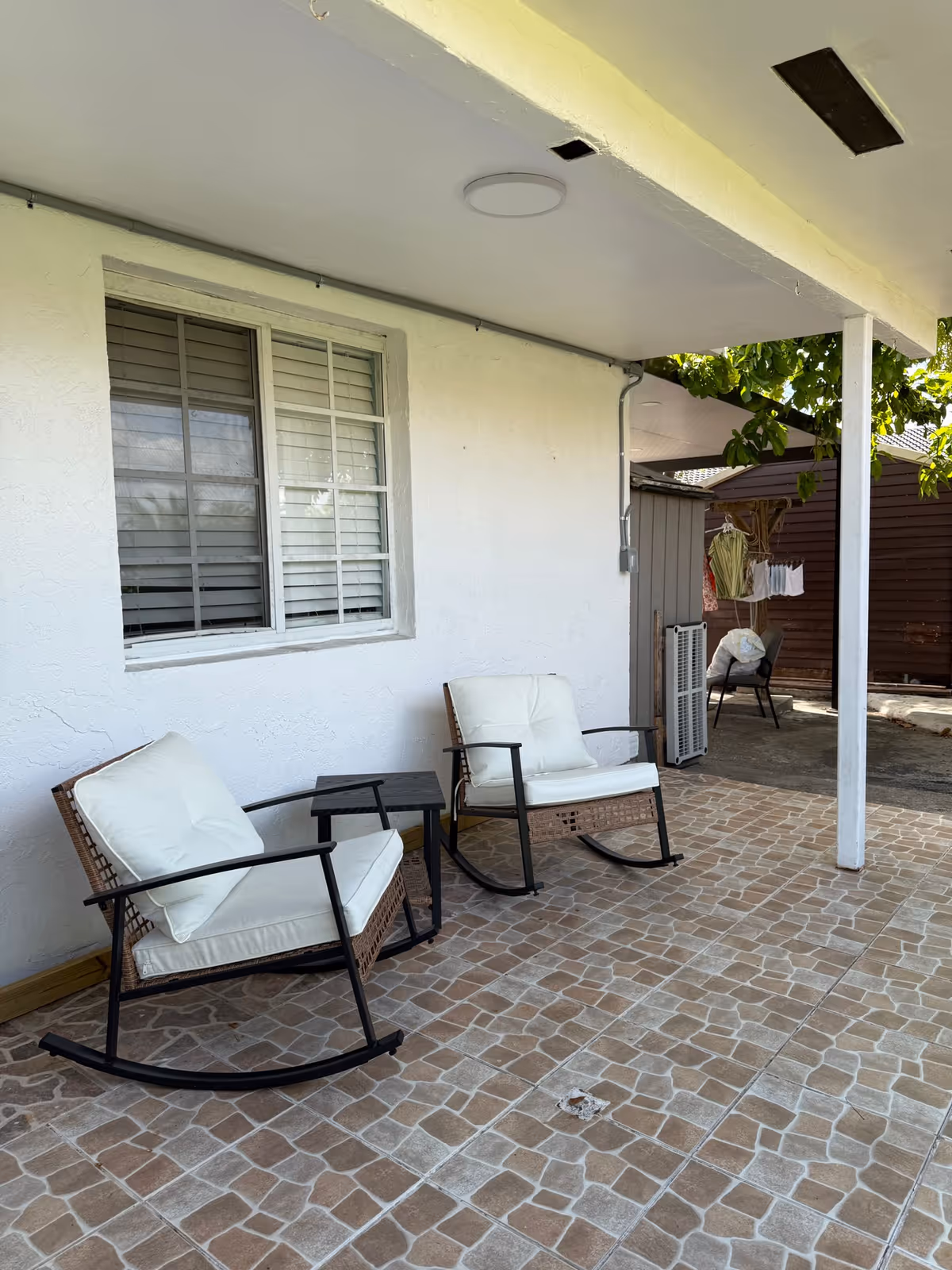 A covered outdoor patio area with two cushioned rocking chairs and a small side table on a tiled floor. A window with closed blinds is on the white wall behind the chairs. In the background, there is a small shed, a chair, and some hanging laundry under a tree.