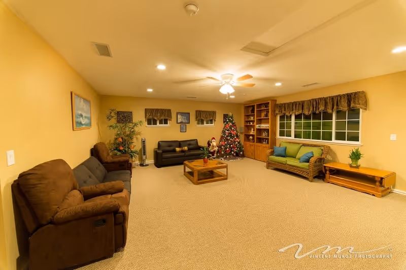 A cozy living room with beige walls and carpet, featuring a brown recliner and sofa on the left, a black leather sofa against the far wall, and a wicker sofa with green cushions on the right. A decorated Christmas tree and a Santa Claus figure are placed near a wooden built-in shelving unit. A wooden coffee table is centered on the carpet, and a ceiling fan with lights is mounted on the ceiling. There are three small windows with valances on the far wall and a larger window with a valance on the right wall. A potted plant and a wooden side table are also visible.