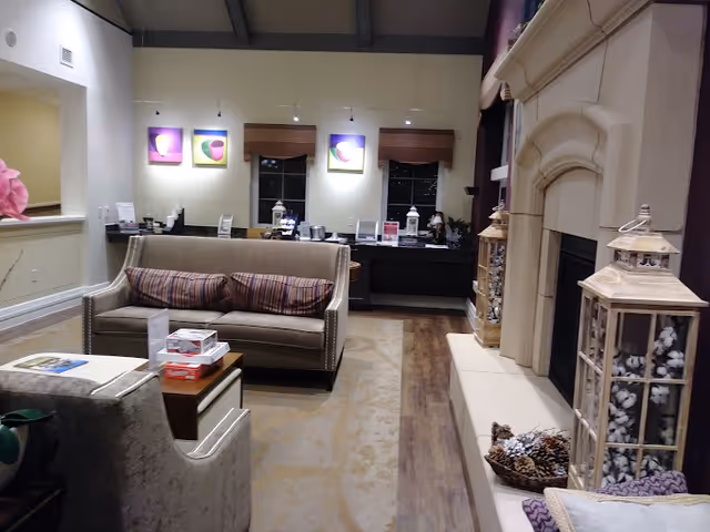 A cozy interior living area featuring a beige sofa with striped cushions, a matching armchair, and a wooden coffee table. The room has a large stone fireplace decorated with lanterns filled with cotton and pinecones. The back wall has three windows with wooden blinds and colorful abstract paintings above them. The floor is a combination of wood and carpet, and the ceiling has exposed beams with hanging lights.