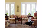 A cozy living room with a floral patterned sofa, a wooden coffee table, two side tables with lamps, a red armchair, and a black dog lying on a rug in front of the sofa. The room has large windows letting in natural light and light yellow walls.