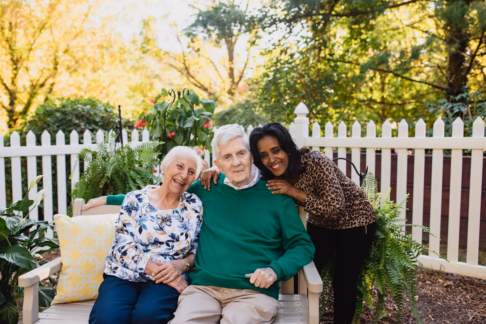 Three elderly individuals sitting and standing closely together on a wooden bench in a garden area with a white picket fence and green plants in the background. They are smiling and appear to be enjoying each other's company on a sunny day.
