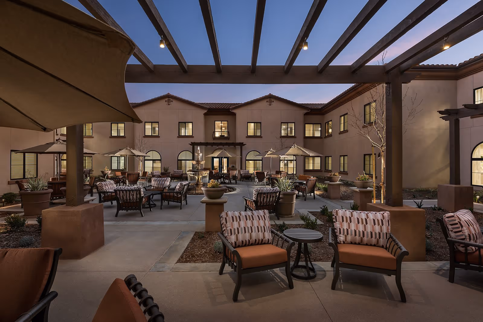 Well-lit central courtyard with patio seating, umbrellas, planters and a fountain surrounded by a two-story building at dusk.