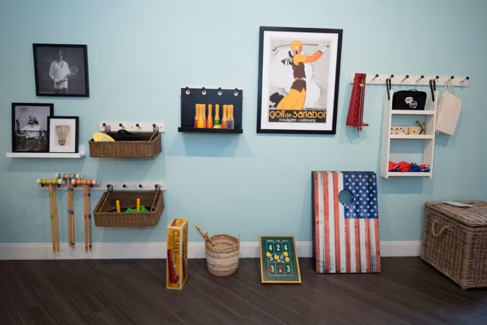 A light blue wall with various shelves and hooks holding vintage-style toys and games including croquet mallets, wooden pins, a bean bag toss board with an American flag design, and framed black and white photos and a colorful poster. There is a wicker basket on the floor and a wicker storage chest to the right.