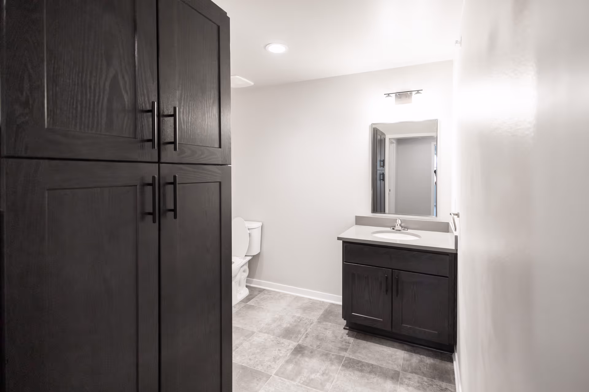 A modern bathroom with dark wood cabinets, a white toilet, a sink with a dark wood vanity, a rectangular mirror above the sink, and a light fixture above the mirror. The floor is tiled in a gray pattern and the walls are painted white.
