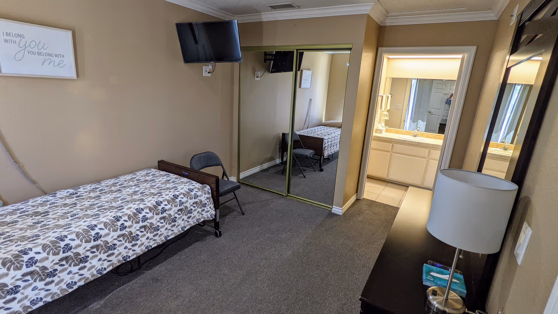 A small bedroom in an assisted living facility with a single bed covered in a patterned bedspread, a folding chair beside the bed, a wall-mounted TV, a large mirror closet with sliding doors, and a dresser with a lamp and tissues. The room leads to a bathroom with a sink and mirror visible.