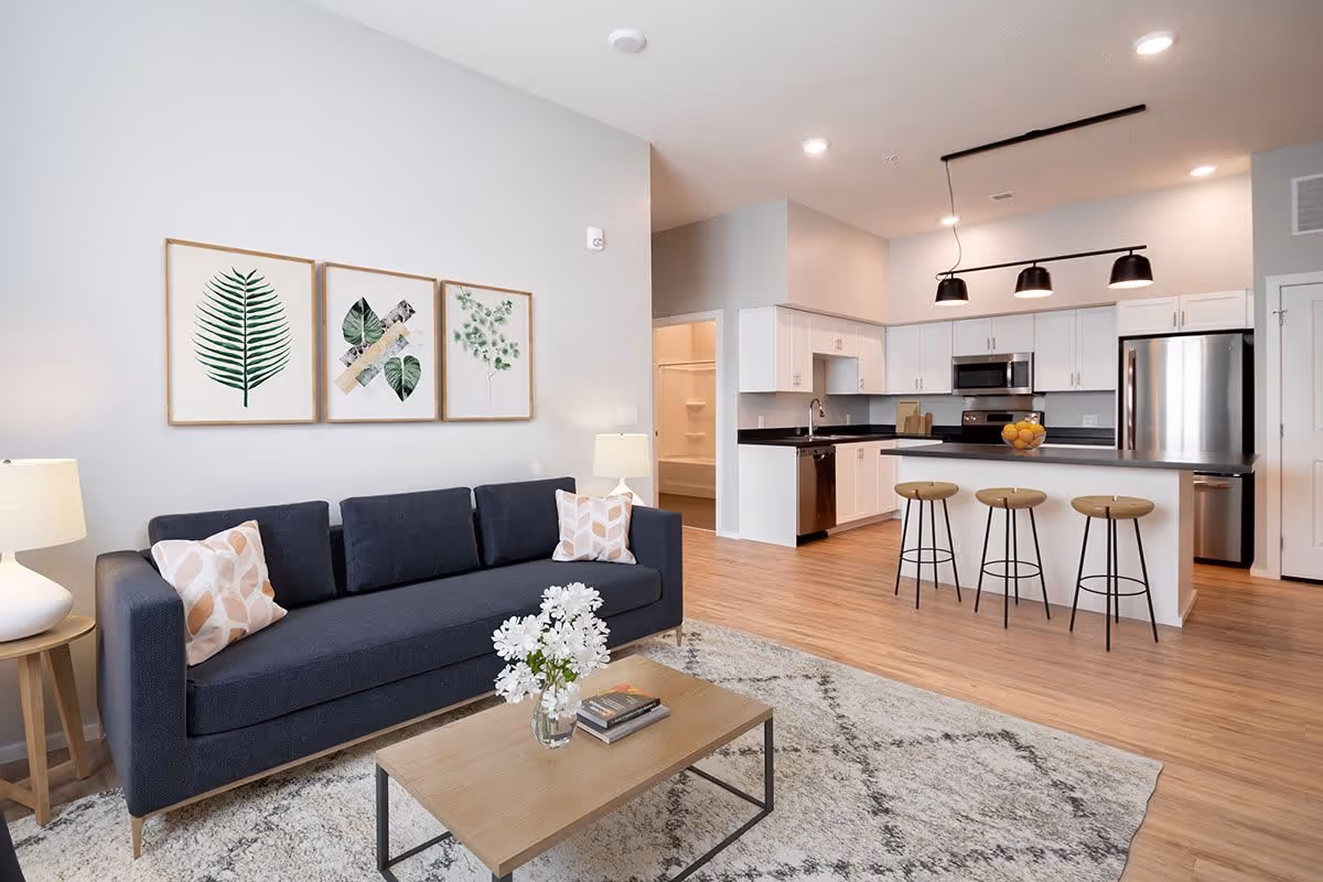 A modern open-concept living room and kitchen area featuring a dark blue sofa with patterned pillows, a wooden coffee table with a vase of white flowers and books, a light-colored area rug, and three framed botanical prints on the wall. The kitchen has white cabinets, black countertops, stainless steel appliances, and a breakfast bar with three wooden stools under black pendant lights.