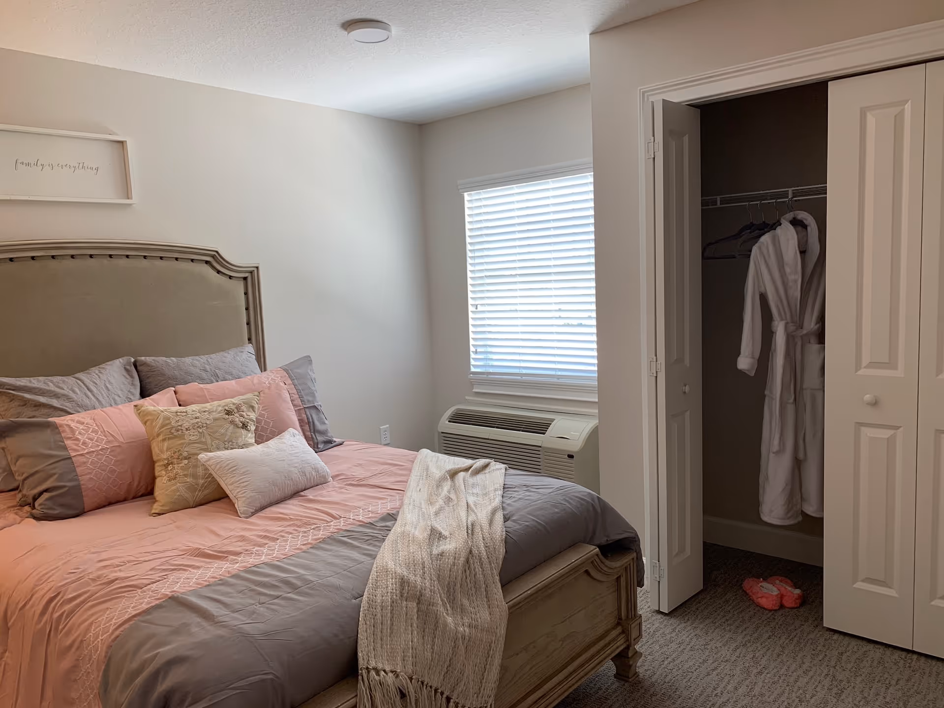 Neatly made bedroom with a pink-and-gray bed, headboard, window AC unit, and an open closet showing a white robe and slippers.