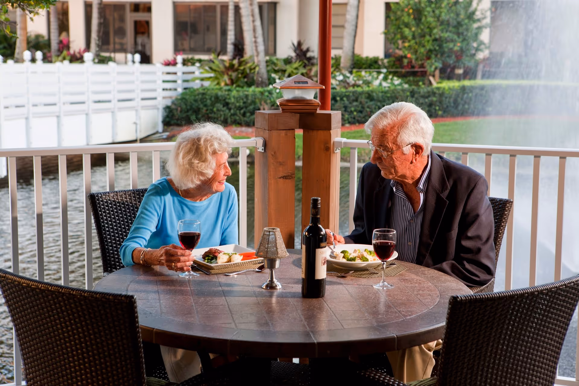 An elderly woman and man sitting at a round outdoor table enjoying a meal and glasses of red wine. They are engaged in conversation with a water fountain and greenery in the background.