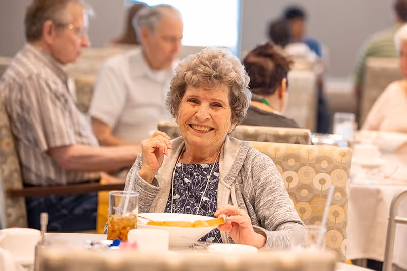 An elderly woman smiling and sitting at a dining table in a senior living facility, holding a bowl of food with a glass of iced tea in front of her. Other elderly people are seated at tables in the background.
