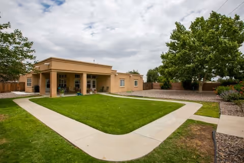 Single-story building with a covered patio area, surrounded by a well-maintained lawn and paved walkways. Trees and shrubs are visible around the property under a partly cloudy sky.