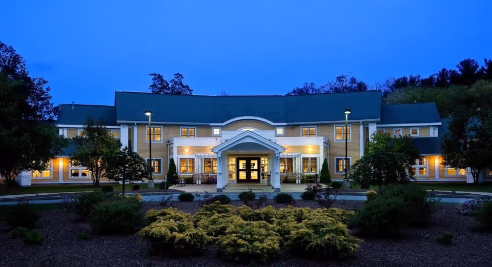 The Arbors of Bedford building exterior at dusk with lights glowing from windows, a covered entrance, and landscaped bushes and trees in front.