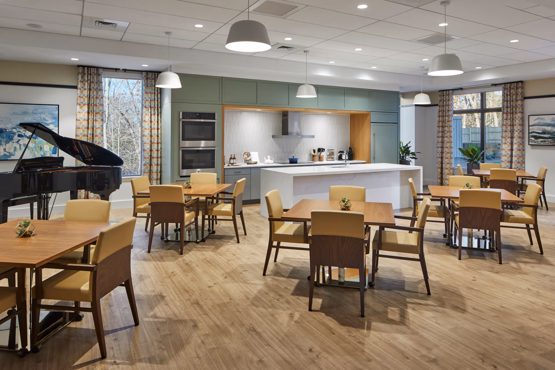 A spacious and modern dining area in a senior living facility featuring several wooden tables with beige cushioned chairs. The room includes a kitchen area with green cabinetry, a white island, and built-in ovens. Large windows with patterned curtains allow natural light to enter. A black grand piano is positioned near one window, and pendant lights hang from the ceiling.