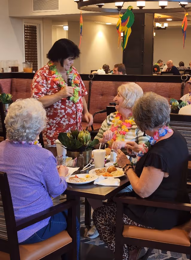 A group of elderly women sitting around a dining table enjoying a meal together in a senior living facility. One woman standing and holding a microphone is engaging with the seated women, who are wearing colorful leis. The room has warm lighting and decorative hanging parrots.