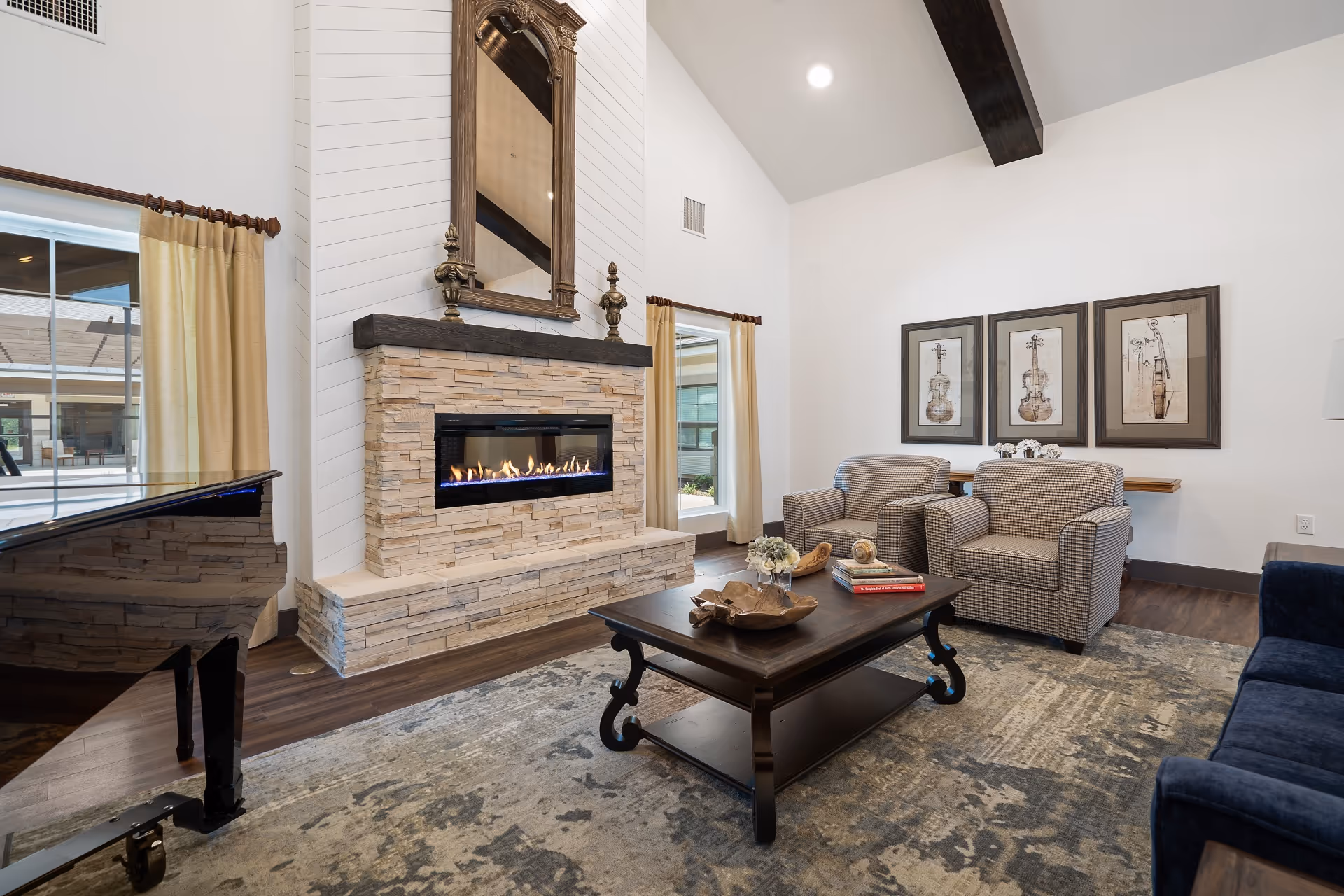 A cozy living room with a modern stone fireplace featuring a lit fire, a large ornate mirror above it, two patterned armchairs, a dark wooden coffee table with decorative items and books, a blue sofa, a grand piano, and three framed violin artwork pieces on the wall. The room has high ceilings with exposed dark wooden beams and large windows with beige curtains.