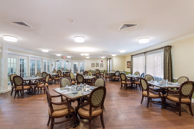 A spacious dining room in an assisted living facility with multiple round tables and cushioned chairs arranged neatly. Each table is set with placemats, napkins, and silverware. The room has wooden flooring, large windows with curtains, and soft ceiling lights providing a warm ambiance.