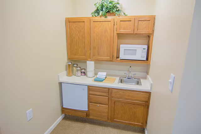 Small kitchenette area with wooden cabinets, a white countertop, a small sink, a microwave, a mini refrigerator, and various kitchen items including jars, paper towels, and folded towels.