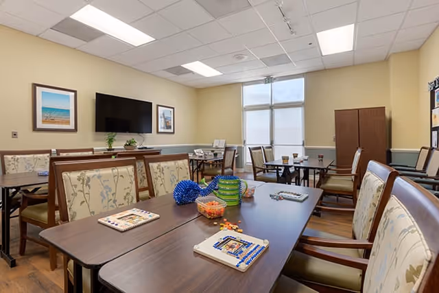 A well-lit room with several tables and chairs arranged for group activities. On the tables are various craft supplies and games. The walls are painted light yellow with framed pictures and a large flat-screen TV mounted on one wall. The room has a window with blinds and a wooden cabinet in the corner.