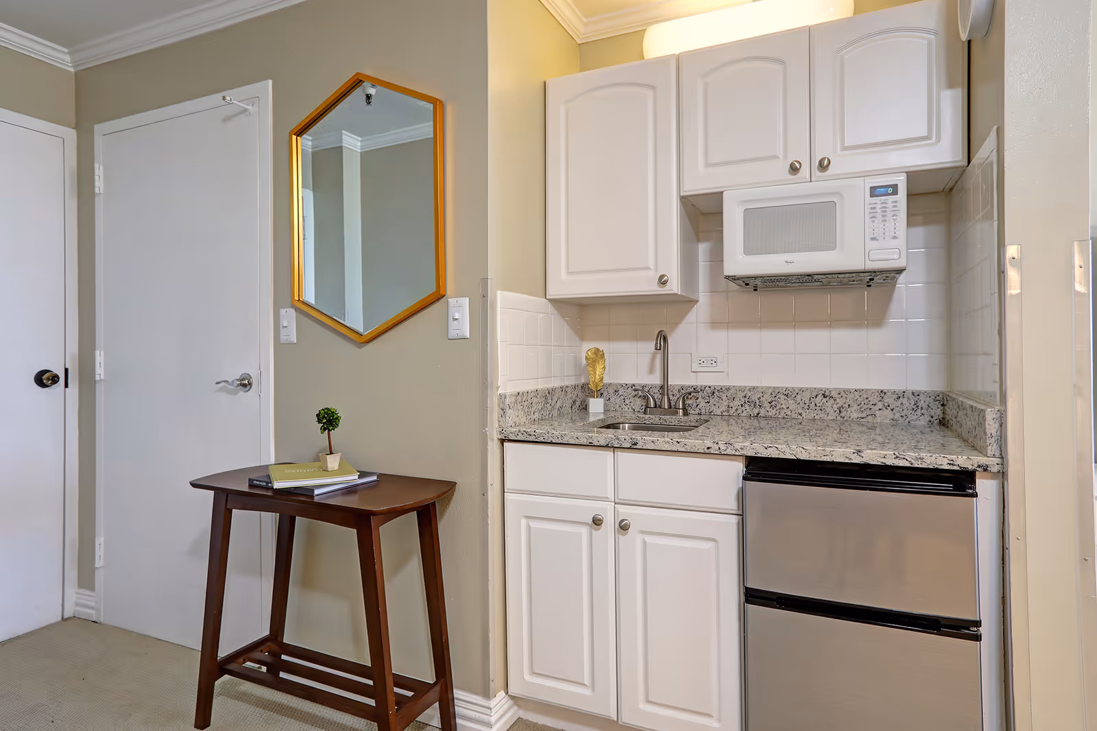 Small kitchenette with white cabinets, granite countertop, a microwave over the sink, a mini fridge, and a small wooden side table beneath a wall mirror.