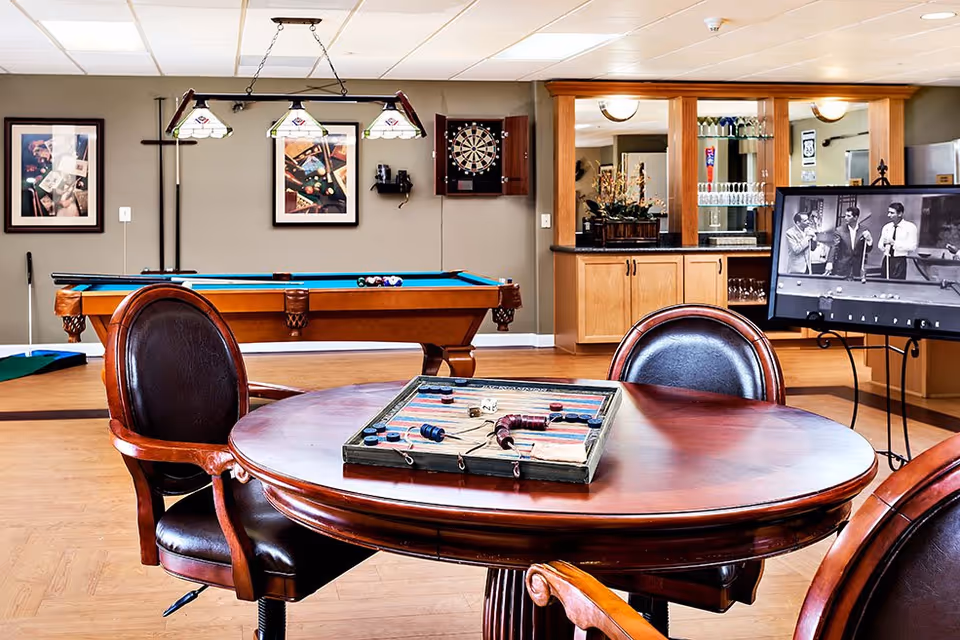 A recreational room featuring a round wooden table with a backgammon game set on it, surrounded by leather chairs. In the background, there is a pool table with a hanging light fixture above it, a dartboard mounted on the wall, framed pictures, and a bar area with glassware and cabinets. A black and white photo is displayed on an easel near the bar.