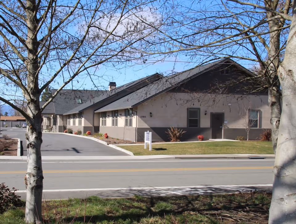 Exterior view of a single-story building with a sloped roof, beige and dark brown walls, and several windows. The building is set behind a road with two leafless trees in the foreground and a small lawn area with some shrubs near the entrance.
