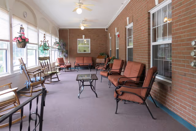 A spacious indoor seating area with brick walls and large windows along one side. The room features several cushioned chairs and sofas arranged around a glass-top coffee table. There are also wooden rocking chairs near the windows, hanging flower baskets, ceiling fans, and potted plants, creating a comfortable and inviting atmosphere.
