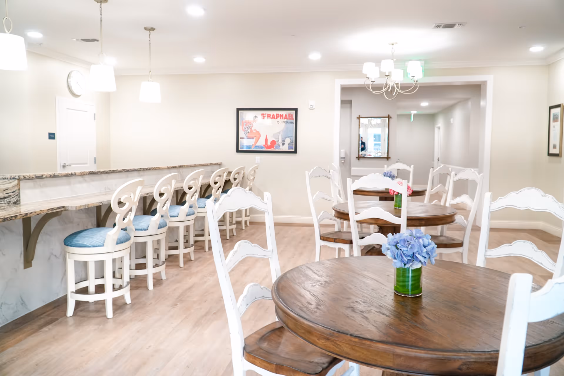 A bright dining area with round wooden tables and white chairs, each table decorated with a small vase of blue flowers. To the left, there is a marble countertop with several white bar stools featuring blue cushions. The walls are light-colored with framed artwork and a clock, and ceiling lights illuminate the space.