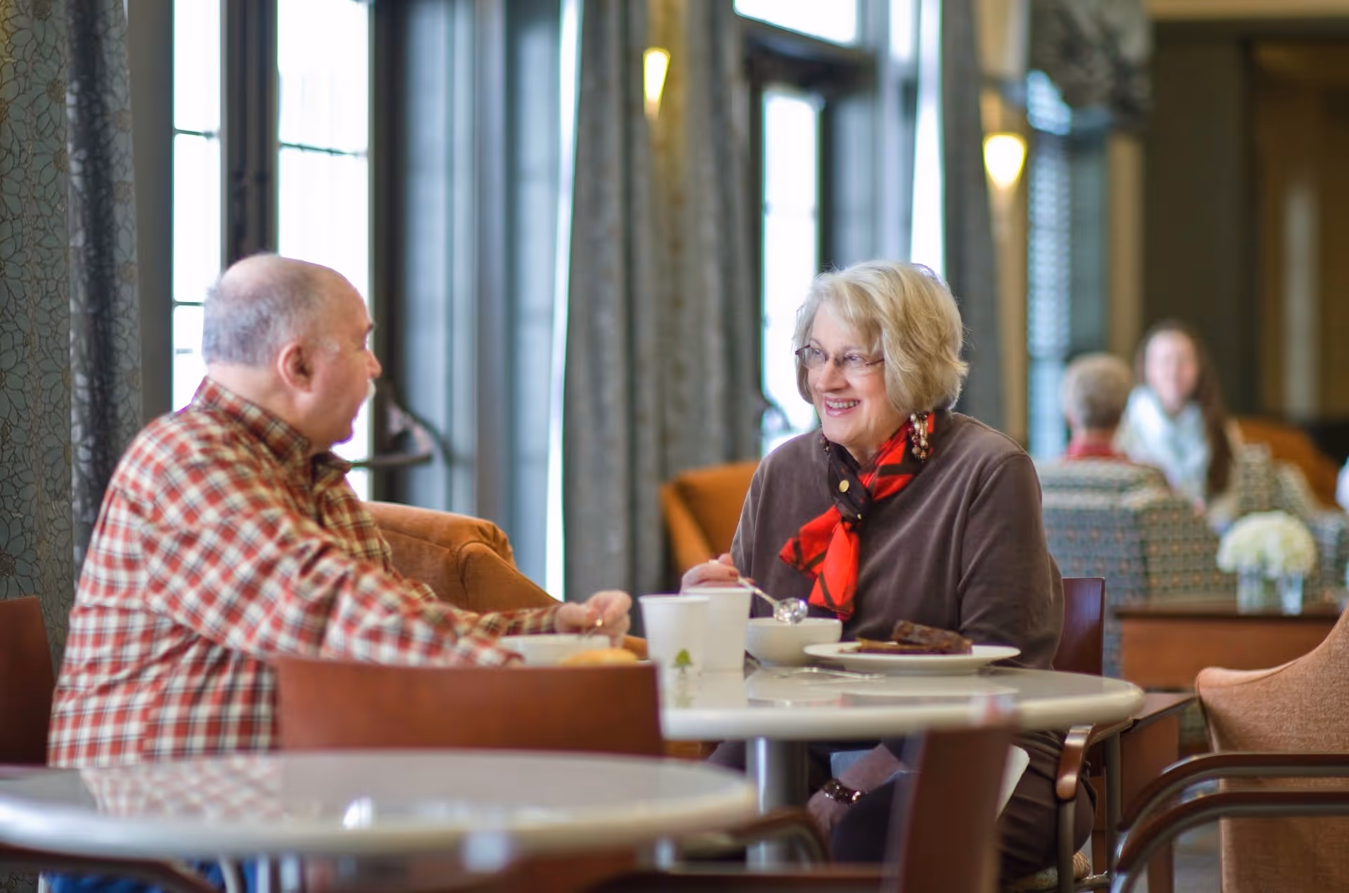 Two older adults sit at a table in a bright communal dining/lounge area, chatting over coffee and dessert.