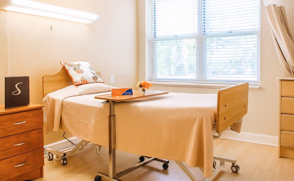 A sunlit nursing facility patient room with an adjustable bed, overbed table, dresser and a large window.