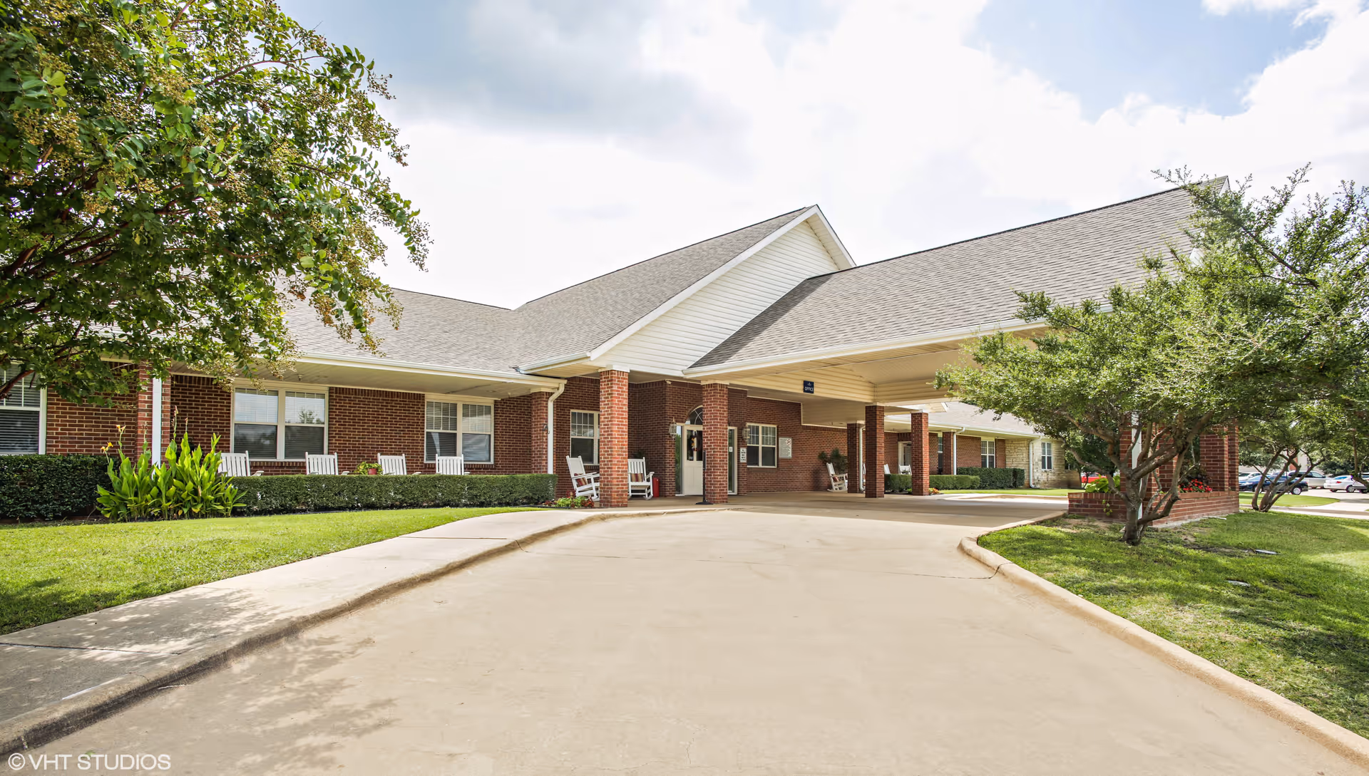 Exterior view of Whitley Place senior living facility showing a single-story brick building with a covered entrance driveway, white rocking chairs on the porch, green shrubs, trees, and a partly cloudy sky.