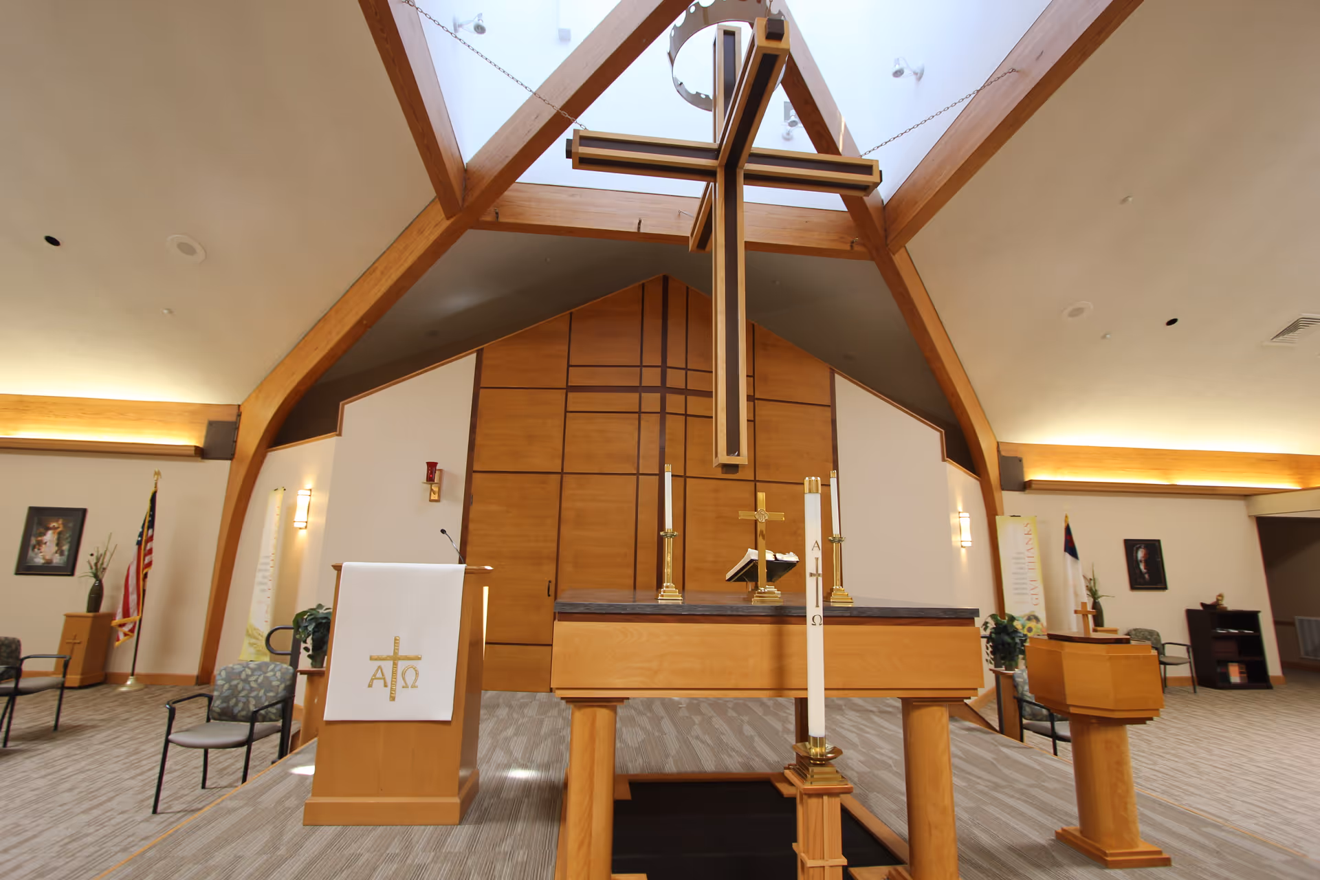Interior view of a chapel with a large wooden cross hanging from the ceiling. The room features wooden beams, a lectern with a white cloth displaying a gold cross and Alpha and Omega symbols, an altar with candles and a Bible, and chairs arranged around the space. The walls are light-colored with wood paneling and there are flags and framed pictures on the side walls.