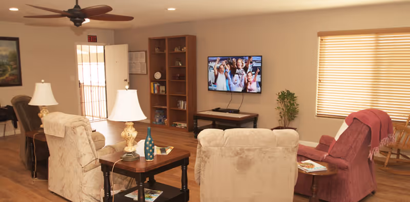 A cozy living room in a senior care facility with several upholstered armchairs arranged around wooden side tables with lamps and decorative items. A flat-screen TV is mounted on the wall showing a group of people smiling. There is a wooden bookshelf with books and decor, a window with blinds, and a ceiling fan above. The room has warm lighting and wooden flooring.
